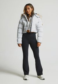 Jauna sieviete, kas ģērbusies īsā baltā pūkainā jaka ar kažokādu, melnās plašās bikses, baltās sporta kurpes un baltu īsu topu, stāv uz baltas fona.