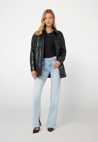 Giubbotto di pelle nera con bottoni a pressione, indossato sopra un top nero e abbinato a jeans azzurri chiari con spacchi laterali. Tacchi a punta neri completano il look.