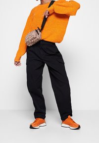Pullover aus grob gestricktem orangefarbenem Material, schwarze Cargo-Hosen mit Seitentaschen und orangefarbene Sportschuhe. Kleine Umhängetasche mit Leopardenmuster und Trageriemen.
