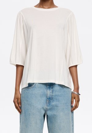 Hvid løs bluse med brede trekvartærmer kombineret med lyseblå højttaljede jeans og et sølvarmbånd på højre håndled.