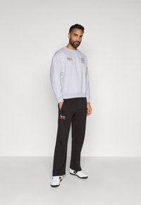 Szürke kereknyakú pulóver logó hímezéssel, fekete melegítőnadrággal, amelyen kiegészítések találhatók, és fehér sportcipővel.