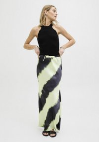 Zwarte geribbelde haltertop gecombineerd met een lange tie-dye rok met zwarte en neon groene strepen. Model draagt zwarte slippers.