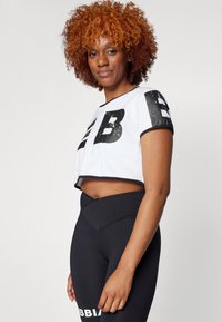 Hvid cropped top med fede sorte bogstaver, parret med højhalsede sorte leggings med et kontrasterende hvidt logo, glat stofstruktur.