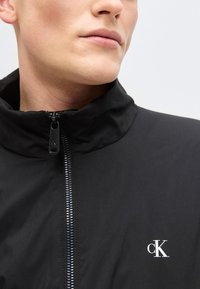 Κοντινό πλάνο ενός ατόμου που φοράει μαύρο μπουφάν με φερμουάρ και μικρό λευκό λογότυπο "cK" στο στήθος, με το φερμουάρ να φτάνει έως το γιακά.