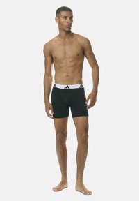 Hombre sin camiseta de pie descalzo, vistiendo bóxers negros con una cinturilla blanca que presenta el logo de Adidas, mirando ligeramente hacia la derecha.