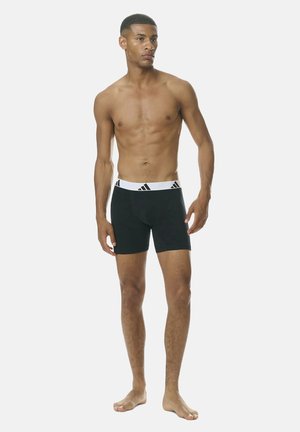 Bovenkledinge man staande op blote voeten, gekleed in zwarte boxershorts met een witte tailleband waarop het Adidas-logo staat, kijkend iets naar rechts.