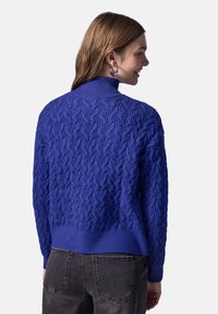 Pull en tricot bleu avec motif en câble, col haut, poignets et ourlet côtelés. Le design présente une finition texturée et une coupe légèrement cropped.