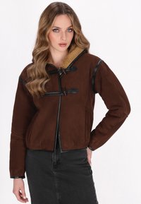 Giacca in suede marrone con colletto in pelliccia shearling, caratterizzata da dettagli in pelle nera e chiusure a toggle. Taglio corto, maniche lunghe e tasche laterali.