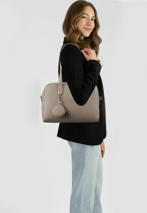 Junge Frau im schwarzen Blazer und hellen Jeans, die eine beiget Lederhandtasche mit rundem Täschchen trägt, leicht lächelnd, vor einfarbigem Hintergrund stehend.