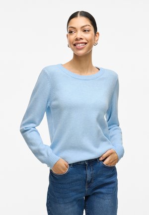 Hellblauer, weich gestrickter Pullover mit rundem Halsausschnitt und gerippten Bündchen. Kombiniert mit blauen Jeans, die Hände in den Taschen.