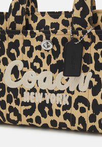 Kangaskassi leopardikuosisena, jossa on beige ja musta kangas, "Coach"-logo brodeerattuna vaalean kerman värisenä, hopeaa jalostusta ja musta tagi.