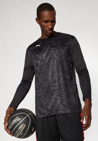 Black long-sleeve athletic shirt featuring a zigzag pattern, made of breathable fabric. Positioned near a black basketball with a logo.