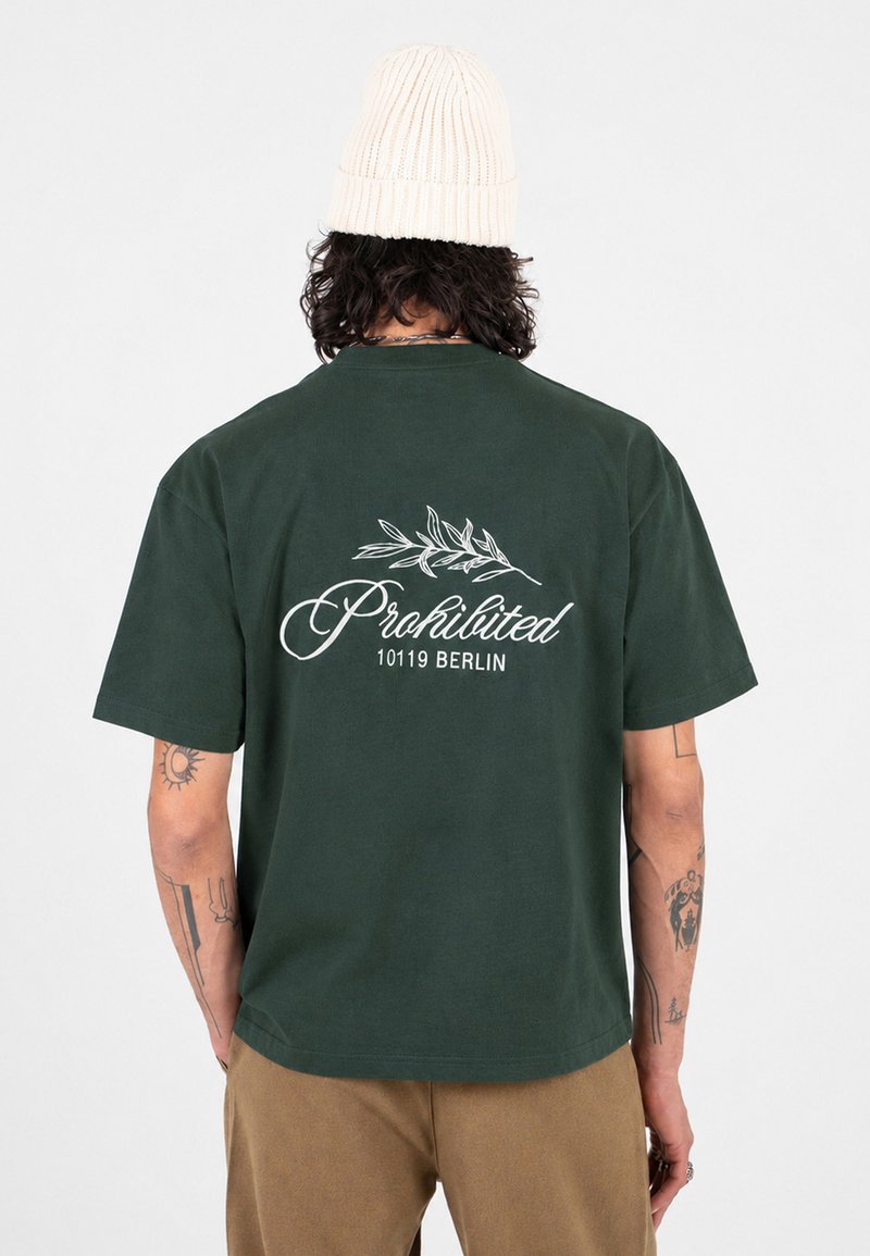 Homme avec des tatouages portant un t-shirt vert foncé avec l'inscription « Prohibited 10119 Berlin » et un motif feuille, un pantalon beige et un bonnet en laine blanc.