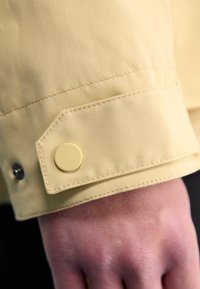 Polsino di giacca gialla con chiusura a bottone tondo, texture liscia e dettagli di cucitura visibili, tenuto sopra una mano contro uno sfondo scuro.