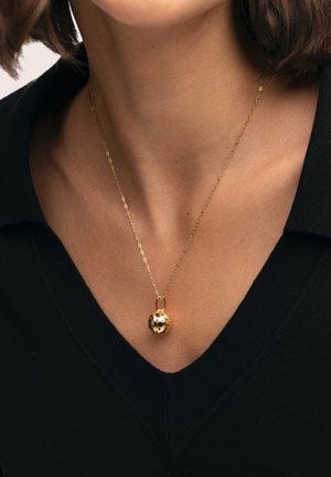 Collar dorado con diseño de eslabones y un colgante esférico con superficie texturizada y pequeñas motas negras, llevado sobre una prenda negra.