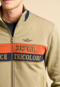 Giacca verde con zip, caratterizzata da strisce orizzontali arancioni e blu navy, con il testo "313° GR." e "TRICOLORI", e un'emblema alato sul petto.
