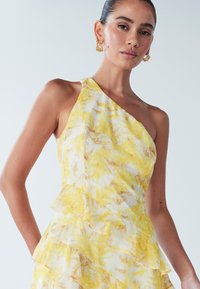 Asimetriska dzeltena kleita ar baltu un gaiši brūnu ziedu rakstu, kas ir ar kārtainām volānu detaļām un gludu, vieglu audumu.