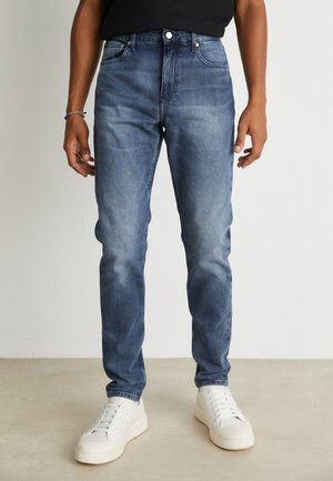 Vaqueros azules con un lavado desgastado, corte ajustado, diseño de cinco bolsillos y cierre de un solo botón, llevados con zapatillas blancas.