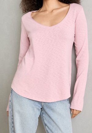 Kobieta ubrana w jasnoróżową bluzkę z długim rękawem i dekoltem w serek oraz jasnoniebieskie dżinsy na neutralnym tle.