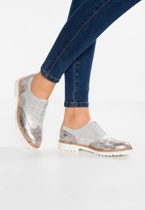 Chaussures à lacets argentées et grises avec un design texturé, comprenant des finitions métalliques et mates, une semelle en caoutchouc à profil épais et des accents décoratifs.