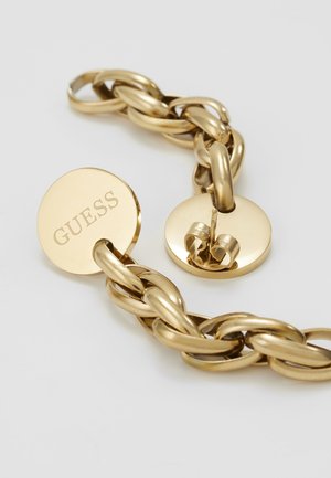 Złota bransoletka łańcuszkowa z grubymi ogniwami i okrągłą zapięciem, jedna strona wygrawerowana logo "GUESS", na prostym białym tle.