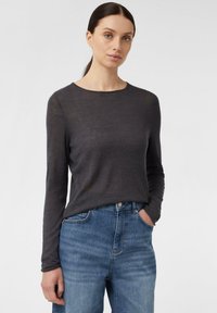 Graues langärmliges Oberteil aus einem weichen Material, mit einem Rundhalsausschnitt. Trägt dazu hellblaue Jeans und hat die Hände in den Taschen.