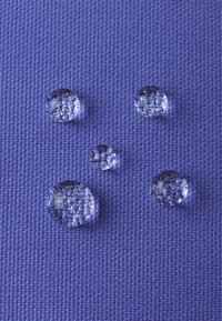 Gotas de agua claras de diferentes tamaños sobre una superficie de tela púrpura texturizada, reflejando la luz y mostrando sus formas lisas y redondeadas.