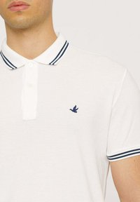 Polo bianco in tessuto strutturato, con strisce blu navy sul colletto e sui polsini delle maniche, e un piccolo logo di un uccello blu navy sul petto.