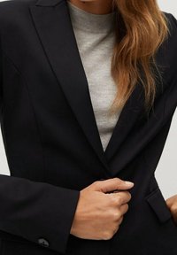 Blazer noir à coupe structurée, fermeture à un bouton, et revers crantés, associé à un pull ajusté gris clair en dessous.