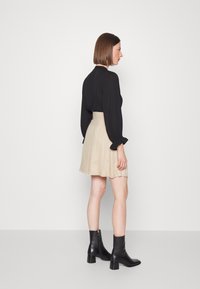 Blusa preta de mangas compridas com punhos franzidos, combinada com uma saia bege. Modelo usa botas pretas de tornozelo com salto robusto. Textura de tecido suave.