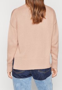 Jasnoróżowy sweter z dzianiny z ribbed hemem, okrągłym dekoltem i opadającymi ramionami, noszony na niebieskich dżinsach z kontrastującymi łatami.