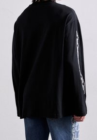 Musta pitkähihainen t-paita, jossa on rento leikkaus, valkoinen graafinen kuvio oikeassa hihassa, valmistettu pehmeästä puuvillamateriaalista.