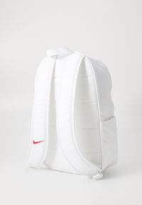 Mochila blanca con correas acolchadas, cierre de cremallera y un logo de swoosh rojo de Nike. Presenta un bolsillo frontal y una tela texturizada.