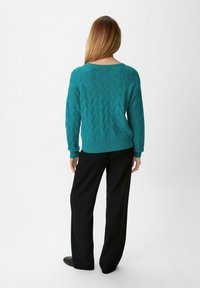 Türkiser gestrickter Pullover mit Zickzackmuster, Rundhalsausschnitt und gerippten Bündchen, kombiniert mit schwarzen weit geschnittenen Hosen.