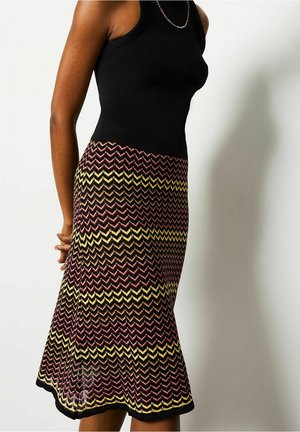 Kobieta w czarnej bluzce bez rękawów i spódnicy dzianinowej we wzór zygzaka w kolorach żółtym, różowym, brązowym i czarnym, stojąca na białym tle.