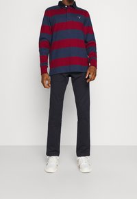Langärmliges PoloheMD in Marineblau mit roten Streifen, geripptem Kragen und Bündchen; kombiniert mit dunklen Hosen und weißen Sneakers.