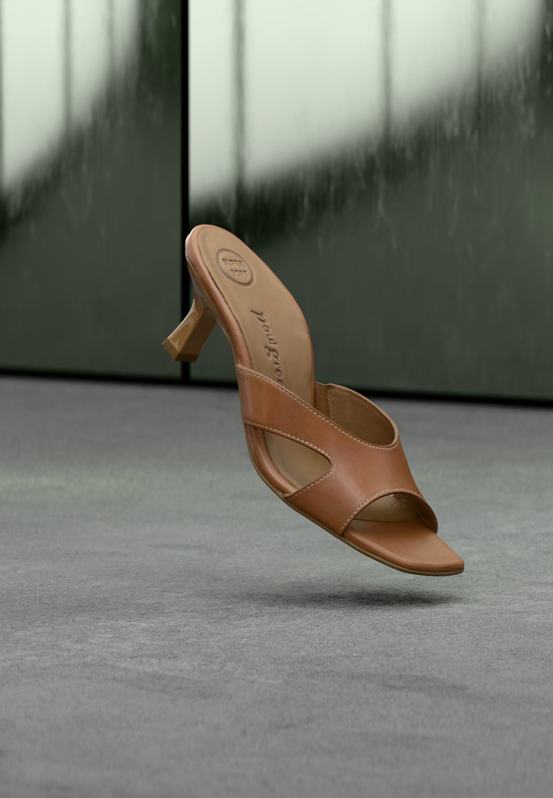 Καφέ δερμάτινο ανοιχτό mule με τακούνι block που αιωρείται πάνω από μια γκρι επιφάνεια, με θολό αφηρημένο φόντο στο πίσω μέρος.