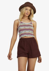 Stribet strik crop top i pastelfarver, parret med løse maroon shorts. Modellen bærer en brun hat; designet har en bølget kant.
