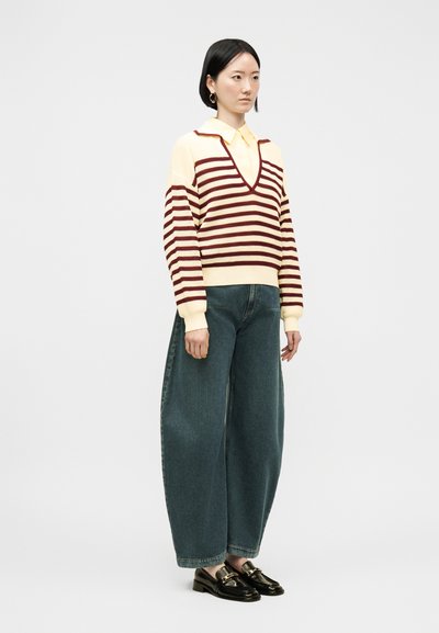 Femme debout portant un pull à col en V rayé crème et bordeaux, une chemise jaune à col, un jean large bleu et des mocassins noirs.
