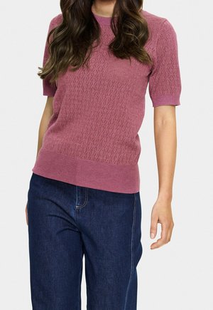Vrouw draagt een roze kort mouw gebreide top met een diamantpatroon, gecombineerd met donkerblauwe spijkerbroek tegen een effen achtergrond.