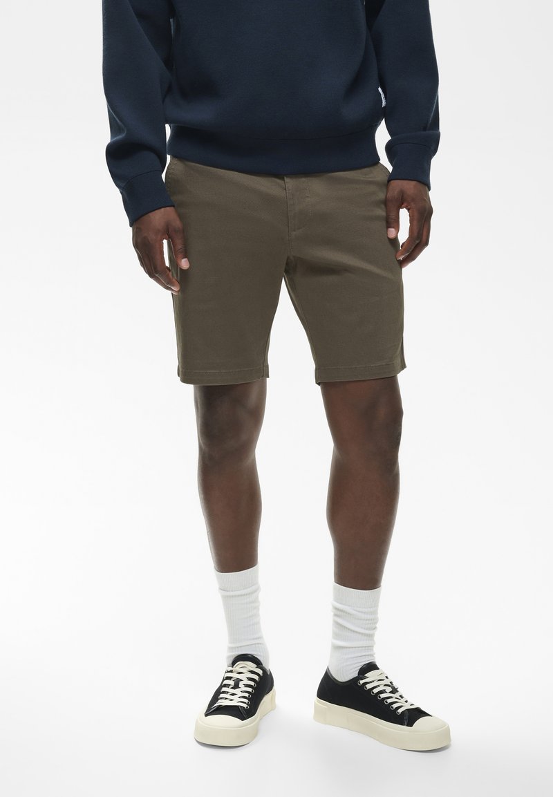Homme portant un short olive, des baskets noires avec des semelles blanches, des chaussettes blanches montantes, et un sweat bleu marine, debout devant un fond uni.
