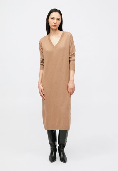 Robe longue en tricot marron avec un col en V et des manches longues. Texture lisse et coupe ample, associée à des bottes noires montantes jusqu'au genou.