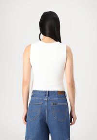 Crop top blanc sans manches associé à un short en denim bleu avec poches arrière et une étiquette en cuir de marque sur la ceinture, texture unie.