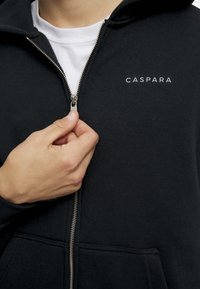 Svart hettejakke med sølvglidelås og kengurulommer, med "CASPARA" i hvit tekst på venstre brystområde. Myk stoffkvalitet.