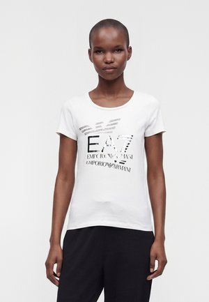 Femme aux cheveux courts portant un t-shirt blanc avec le logo noir EA7 Emporio Armani et un pantalon noir, debout devant un fond uni.