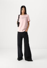 Lyserosa bomull T-skjorte med "TOMMY HILFIGER EST. 1985" trykt i svart, kombinert med vide svarte jeans og en svart skulderveske.