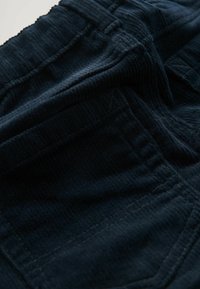 Námornícky kordúrový materiál s vertikálnym ryhovaním. Obsahuje prešité švy a vrecká. Mäkká textúra s matným povrchom.