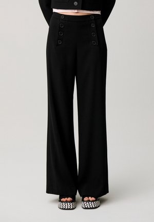 Pantalones negros de talle alto y pierna ancha con doble fila de botones en el frente, combinados con sandalias de plataforma negras y blancas con estampado.