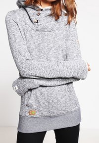 Huppari harmaata sävyä, valmistettu teksturoidusta kankaasta, jossa on huppu, puunapit ja värillinen logopatch alaosassa.