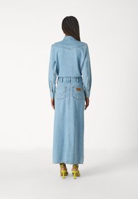 Világoskék farmer hosszú ingruha hosszú ujjal, gombos mandzsettával és két hátsó zsebbel, amelyek zigzag varrással díszítettek. Zöld magassarkú szandál.